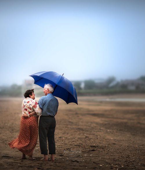 A photograph by Sujata Setia. A couple stands under a blue umbrella, holding each other, in a field.
