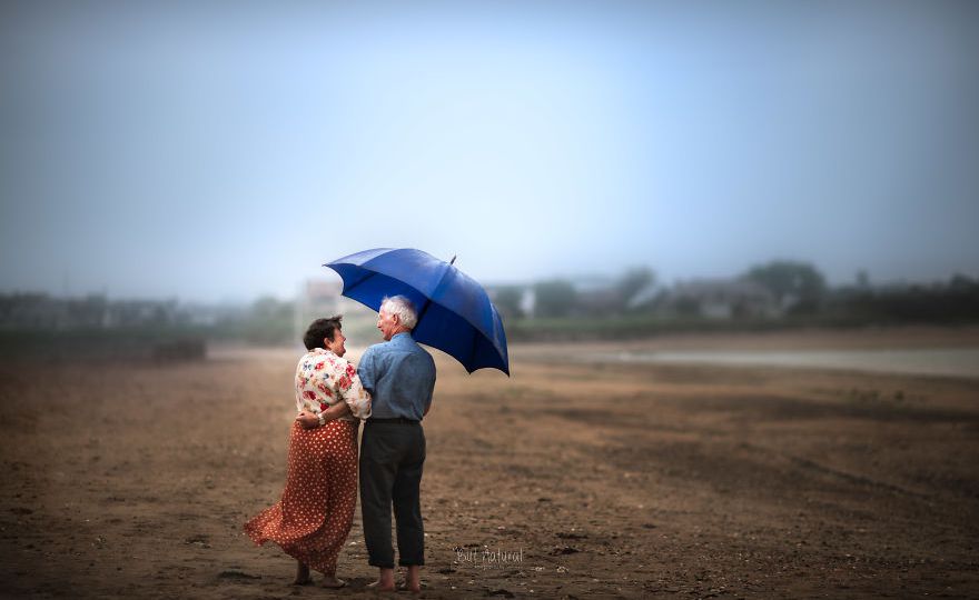 A photograph by Sujata Setia. A couple stands under a blue umbrella, holding each other, in a field.