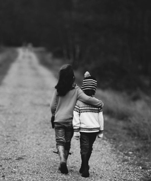 A black and white photograph of two children walking on an empty road. One child’s hand is on the shoulders of the other child