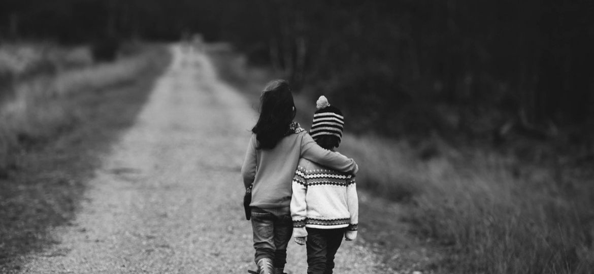 A black and white photograph of two children walking on an empty road. One child’s hand is on the shoulders of the other child