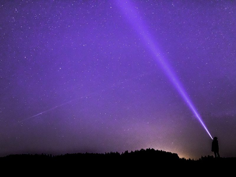 An image of a starry purple sky and a person flashing a white light towards the sky.