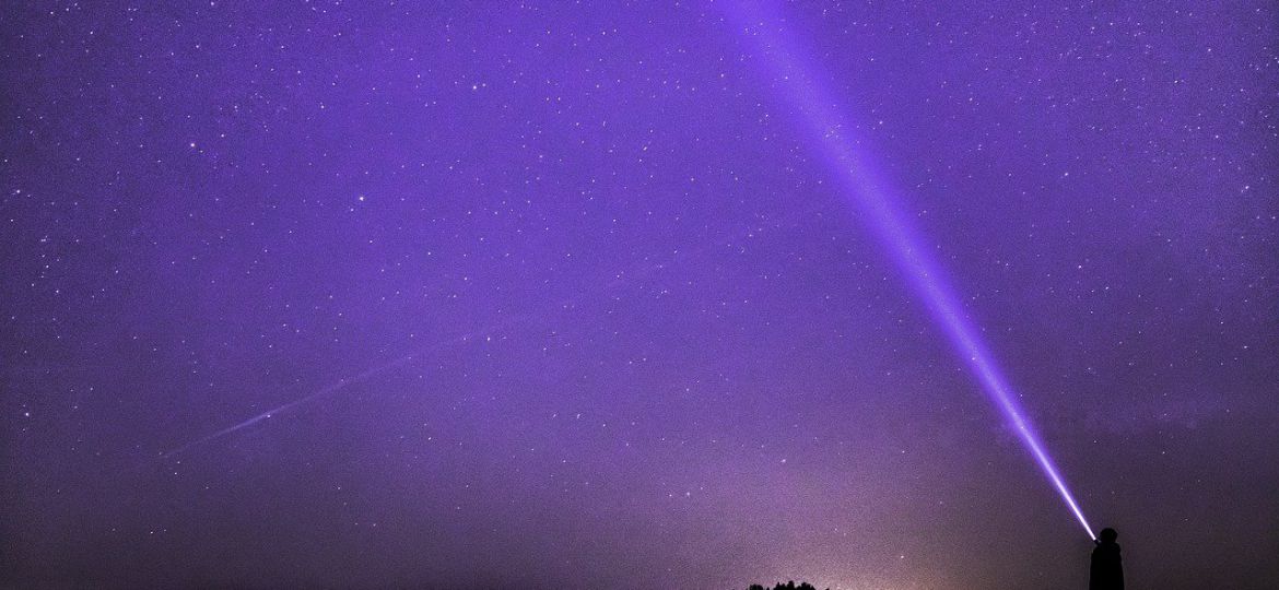 An image of a starry purple sky and a person flashing a white light towards the sky.