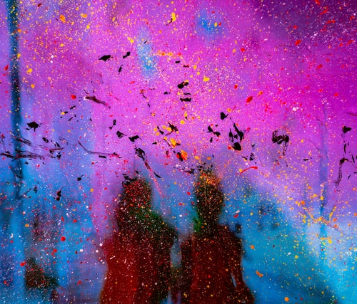 An abstractly painted image with magenta and blue and splashes of red, yellow, and black colour. In the background, two brown-black silhouettes.