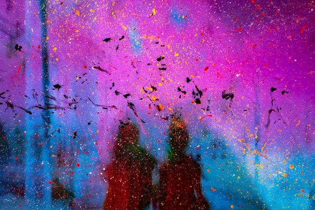 An abstractly painted image with magenta and blue and splashes of red, yellow, and black colour. In the background, two brown-black silhouettes.