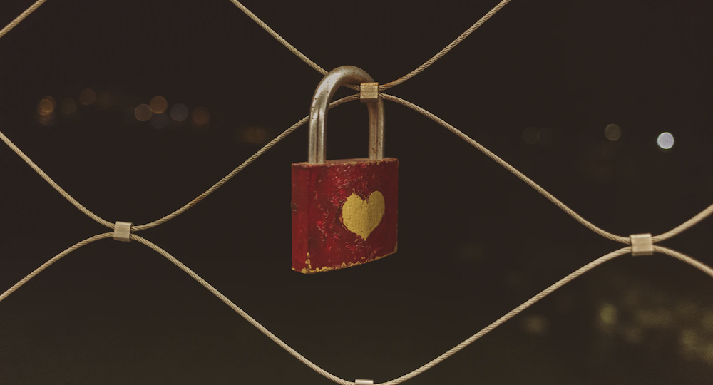 A photograph, against a dark background, of a zoomed-in fence with a square lock hanging. The lock is painted red, left un-coloured in a heart shape in the middle.