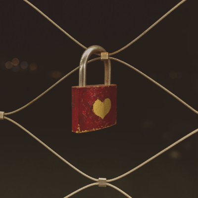 A photograph, against a dark background, of a zoomed-in fence with a square lock hanging. The lock is painted red, left un-coloured in a heart shape in the middle.