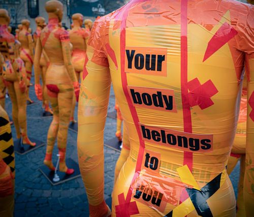 Image of mannequins with orange and pink ductapes. On one of the mannequins ‘Your body belongs to you’ is written