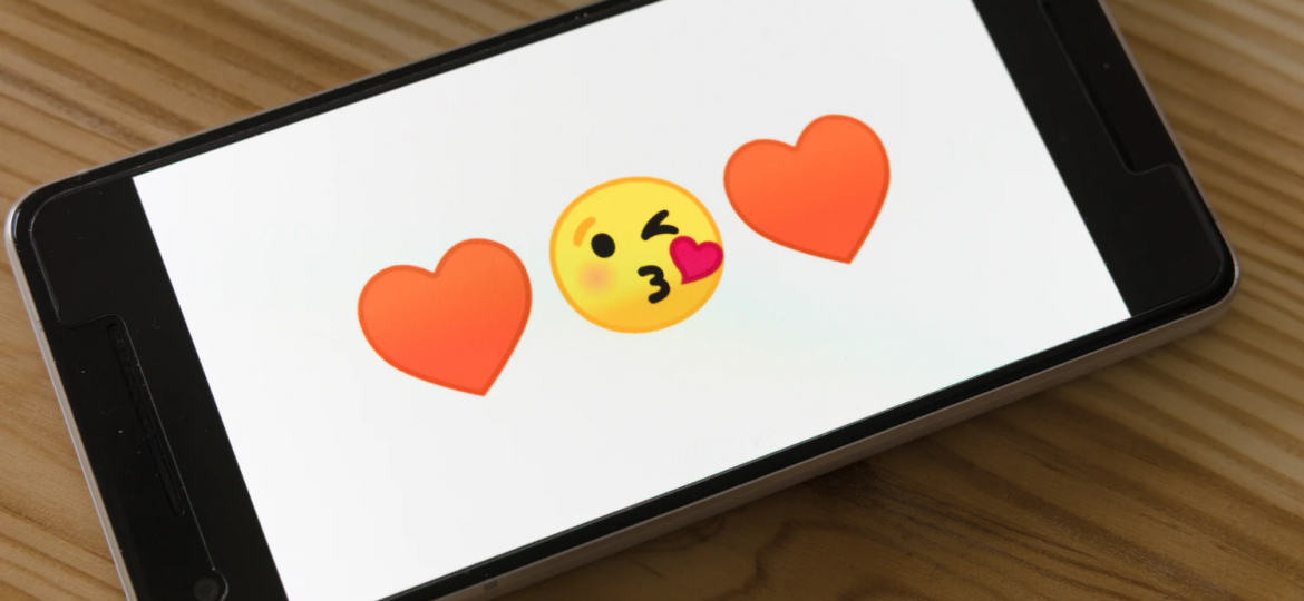 A photograph of a smartphone on a wooden surface. The sides of the phone are black, and the display has a white background with a kissing-face emoticon in the centre and two red heart emoticons on either side.