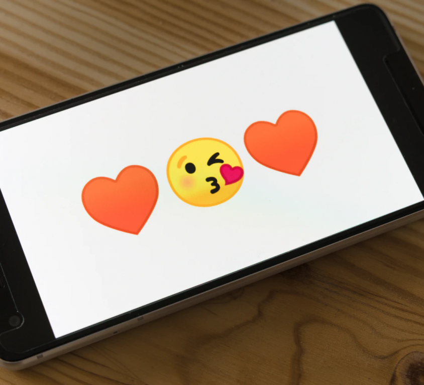 A photograph of a smartphone on a wooden surface. The sides of the phone are black, and the display has a white background with a kissing-face emoticon in the centre and two red heart emoticons on either side.