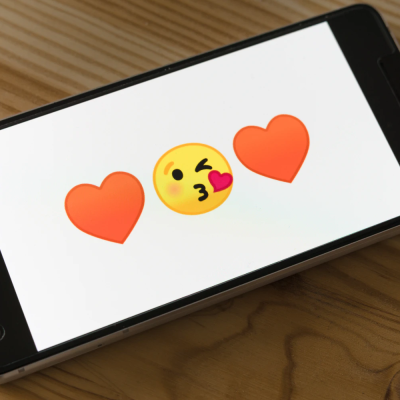 A photograph of a smartphone on a wooden surface. The sides of the phone are black, and the display has a white background with a kissing-face emoticon in the centre and two red heart emoticons on either side.