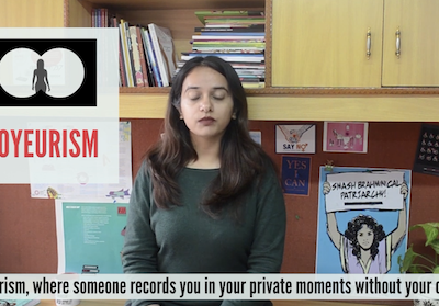 A screenshot of Feminism in India's video How Can Women & Non-Binary People Combat Online Violence? In the middle is Japleen Pasricha, the founder of Feminism in India. She is wearing a grey-black full-sleeved shirt and her eyes are closed. She has deep brown hair reaching under her shoulders on both sides. Behind her is a wooden cupboard stacked with books, and a poster in blue with a woman holding a sign saying 'SMASH BRAHMINICAL PATRIARCHY'. On her left is an illustration that says, in pink, VOYEURISM and above it, an illustration with white circles on black to denote binoculars and a woman's silhouette at the centre. At the bottom of the screenshot are the subtitles: "(a)nd voyeurism, where someone records you in your private moments without your consent."