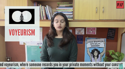 A screenshot of Feminism in India's video How Can Women & Non-Binary People Combat Online Violence? In the middle is Japleen Pasricha, the founder of Feminism in India. She is wearing a grey-black full-sleeved shirt and her eyes are closed. She has deep brown hair reaching under her shoulders on both sides. Behind her is a wooden cupboard stacked with books, and a poster in blue with a woman holding a sign saying 'SMASH BRAHMINICAL PATRIARCHY'. On her left is an illustration that says, in pink, VOYEURISM and above it, an illustration with white circles on black to denote binoculars and a woman's silhouette at the centre. At the bottom of the screenshot are the subtitles: "(a)nd voyeurism, where someone records you in your private moments without your consent."