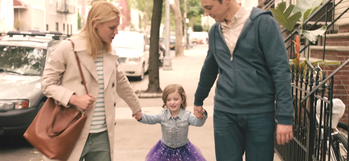 A still from the film 'A Kid Like Jake'. In a city-background with houses to the right and a car to the left, three figures: a blonde woman wearing a black and white striped t-shirt, blue jeans, and a beige coat, carrying a brown bag on her shoulder is holding a child's hand from the left. The child is wearing a purple, glittery tutu and a blue shirt. Their arms are outstretched and on the right side, held by a man wearing a peach shirt, blue hoodie and blue jeans. He has black hair. Both the man and the woman are looking at the child.