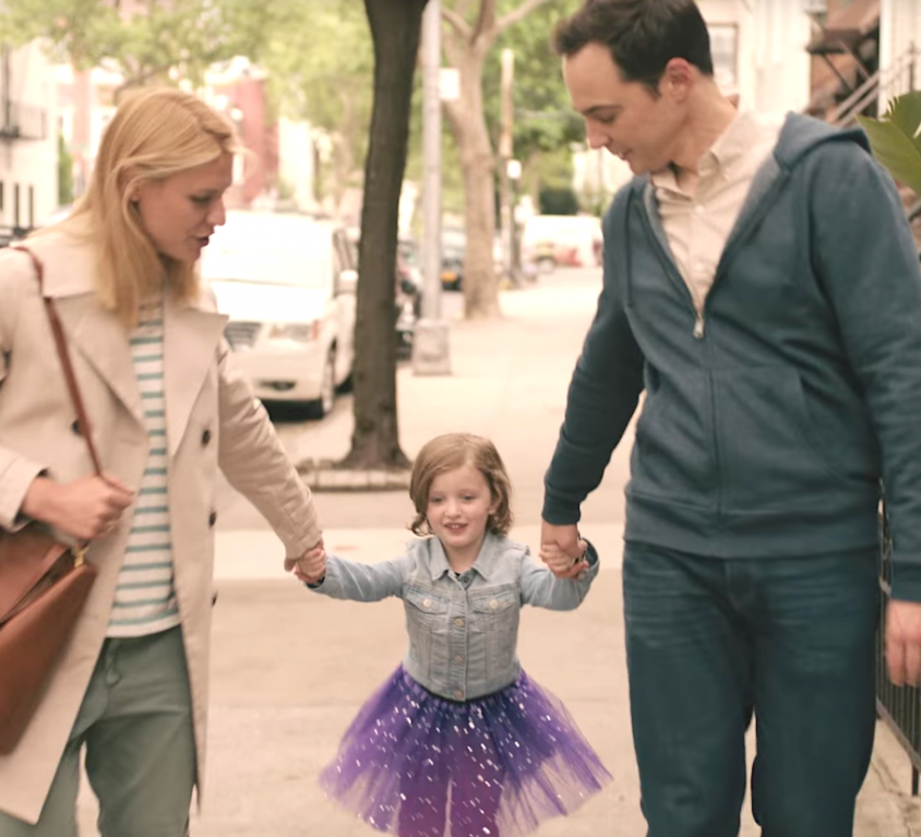 A still from the film 'A Kid Like Jake'. In a city-background with houses to the right and a car to the left, three figures: a blonde woman wearing a black and white striped t-shirt, blue jeans, and a beige coat, carrying a brown bag on her shoulder is holding a child's hand from the left. The child is wearing a purple, glittery tutu and a blue shirt. Their arms are outstretched and on the right side, held by a man wearing a peach shirt, blue hoodie and blue jeans. He has black hair. Both the man and the woman are looking at the child.