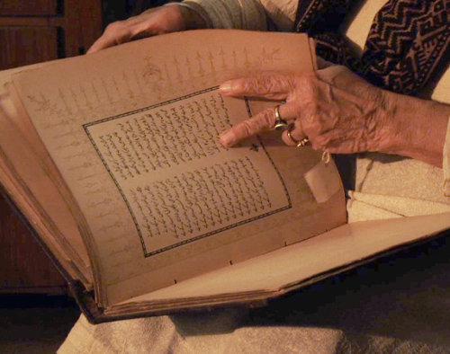 A photograph of an individual with a book. Their face is not visible. They are flipping through a seemingly old book, and are turning the page using two fingers. They are wearing a cream top with a brown scarf and a ring on their finger.