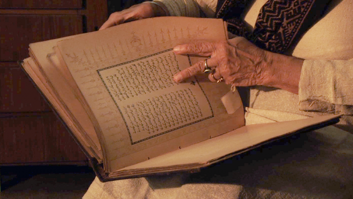 A photograph of an individual with a book. Their face is not visible. They are flipping through a seemingly old book, and are turning the page using two fingers. They are wearing a cream top with a brown scarf and a ring on their finger.