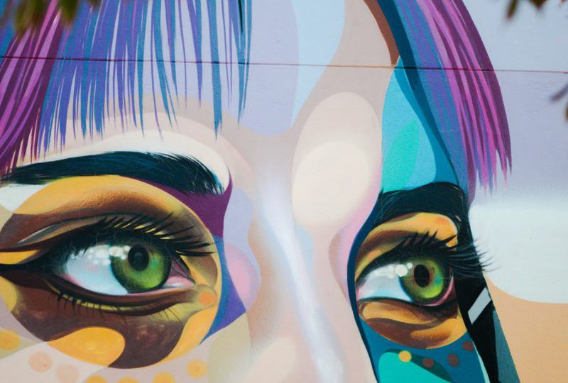Wall-art of a face till the bridge of the nose. The face is coloured in peach, grey, and shades of blue. The eyes, green, are lined in black and have spots of yellow, brown, and blue colour around them. Under the left eye, there are yellow dots. The hair is streaked with purple and blue.