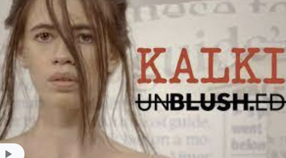 A screenshot of Kalki Koechlin's rendition of 'The Printing Machine'. The background has a fade-effect of newspaper headlines. To the left is Kalki's face till the beginning of her shoulders. She is looking directly at the camera. Her hair is tied with strands across her face. To the left, in big red lettering: KALKI an underneath it, in black and strikethrough: UNBLUSHED. 'BLUSH' is bold.