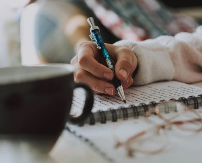 A photograph of a person's arm and hand. The background of a couch with a blue duvet is blurred, and the focus is on the person's hands, with brown nail-paint, and a blue pen in their hands. They are writing in a spiral bound notebook. Beside them is a black cup.