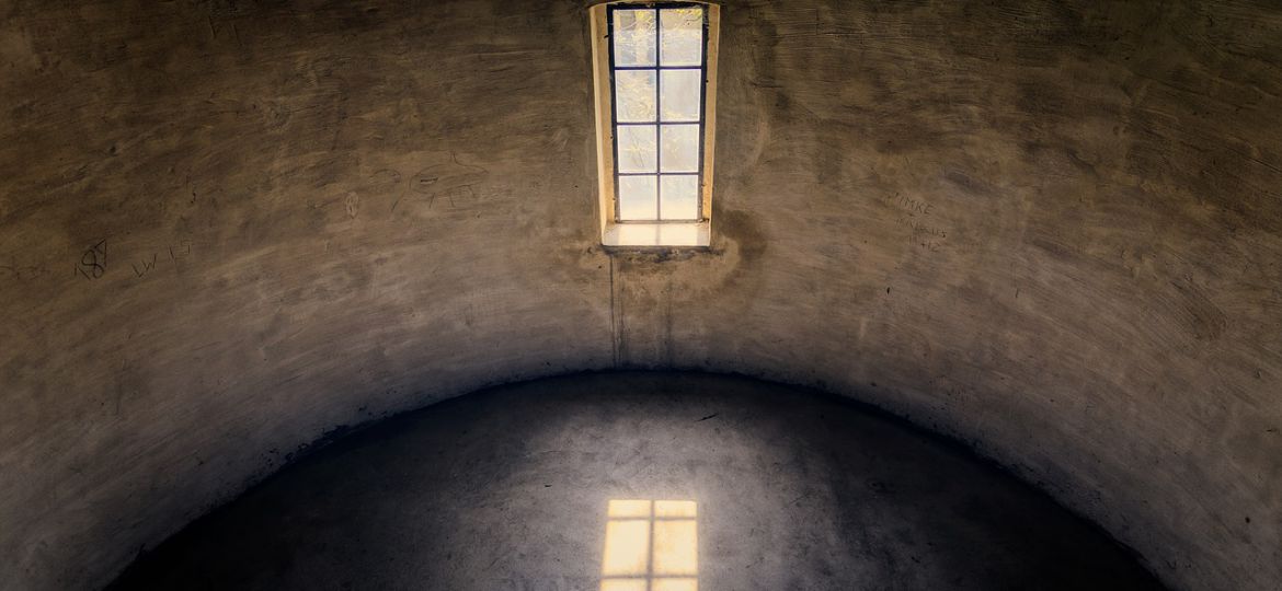 A photograph of a room with one window. The photo showcases the room as semicircular and has a black floor with a window reflected, and brown-white brick walls with a sunlit window.