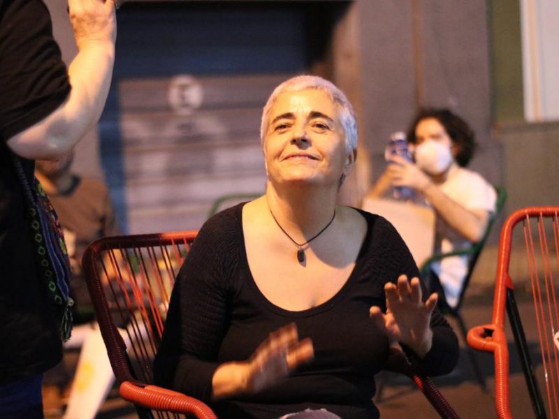 A photograph of a woman sitting indoors. She has cropped white-grey hair and is looking upwards. She is wearing a black full-sleeved top and her palms are open outwards. She is wearing a black necklace and is smiling. In front of her, a person is half-visible, clad in black, holding out their palms. There are red chairs on the side, and at the back, a person with black short hair, wearing a white top and mask, using a phone, is sitting.