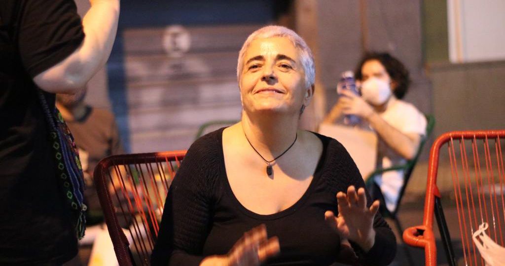 A photograph of a woman sitting indoors. She has cropped white-grey hair and is looking upwards. She is wearing a black full-sleeved top and her palms are open outwards. She is wearing a black necklace and is smiling. In front of her, a person is half-visible, clad in black, holding out their palms. There are red chairs on the side, and at the back, a person with black short hair, wearing a white top and mask, using a phone, is sitting.