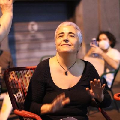 A photograph of a woman sitting indoors. She has cropped white-grey hair and is looking upwards. She is wearing a black full-sleeved top and her palms are open outwards. She is wearing a black necklace and is smiling. In front of her, a person is half-visible, clad in black, holding out their palms. There are red chairs on the side, and at the back, a person with black short hair, wearing a white top and mask, using a phone, is sitting.