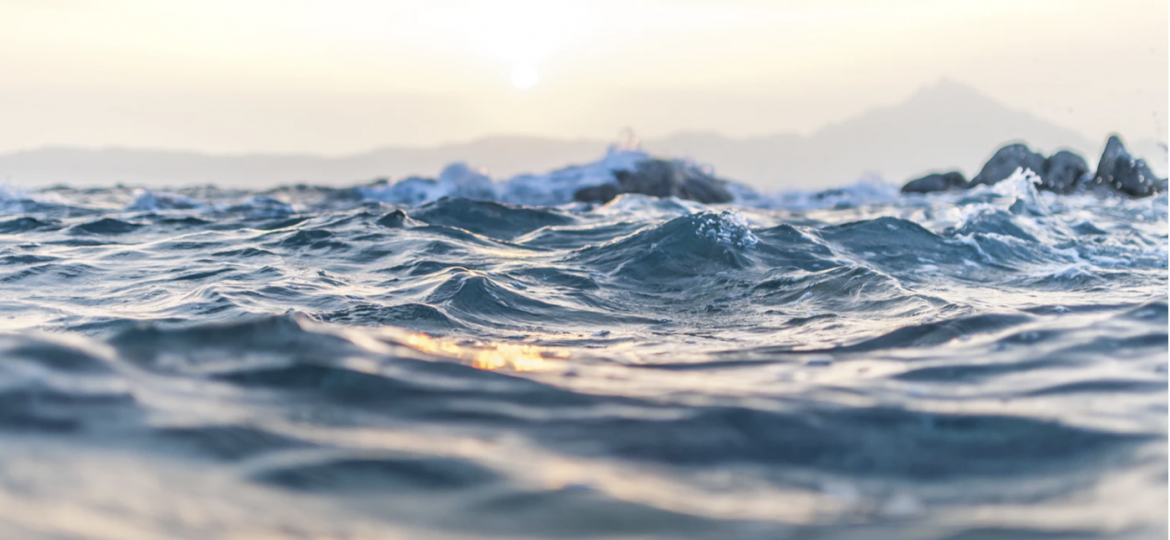 A photograph of a water body. The sky is tinged with pink-mauve-yellow and is reflecting yellow on the different shades of blue of the waves.