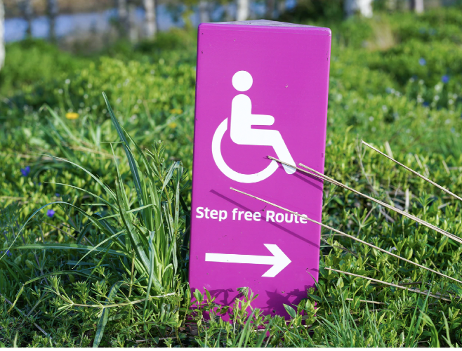 A photograph of a purple-coloured board on grass. The board, in white, has a symbol for a person using a wheelchair and underneath it, the typography: Step free Route. Below it is an arrow pointing to the right.