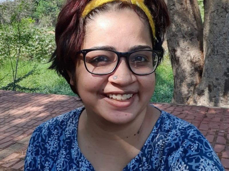 A photograph of disability rights activist Srinidhi Raghavan. She is wearing a blue top with a white pattern, black-rimmed spectacles, and a yellow hair-band.