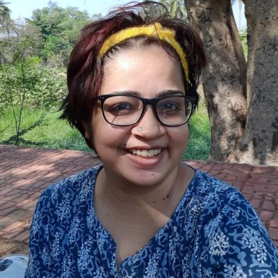 A photograph of disability rights activist Srinidhi Raghavan. She is wearing a blue top with a white pattern, black-rimmed spectacles, and a yellow hair-band.