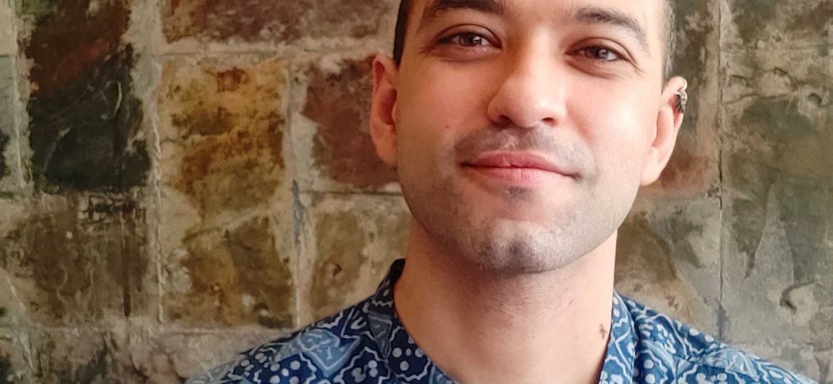 Photo of a smiling man who is wearing a blue patterned shirt and standing in front of a brick wall