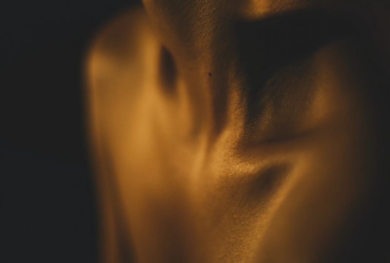 A photo of the throat, clavicles and shoulders against a black background. The body is highlighted with golden brown light and there is a mole at the base of the throat.