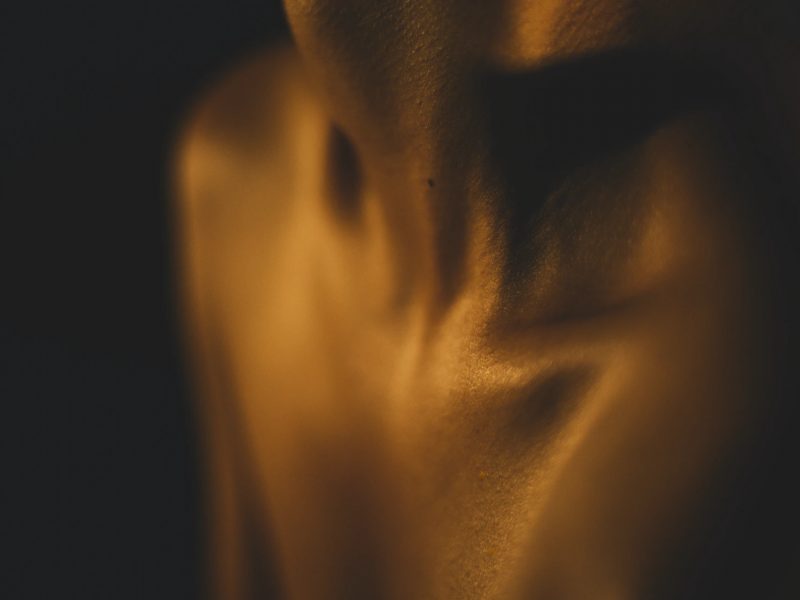 A photo of the throat, clavicles and shoulders against a black background. The body is highlighted with golden brown light and there is a mole at the base of the throat.