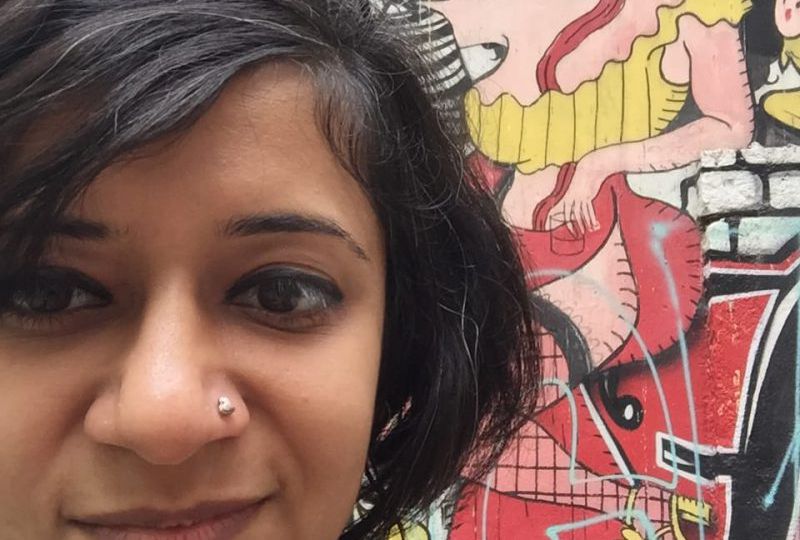 A photograph of a woman’s face with a mural in the background. The woman has short black hair and a nose piercing. She is smiling.