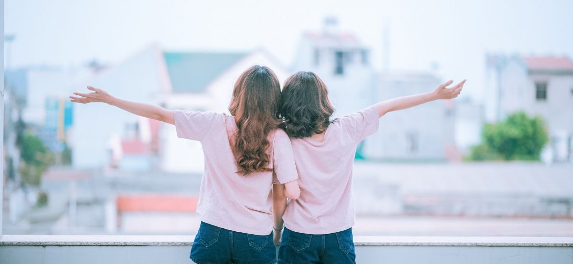 A photograph featuring two women with their backs to the camera and their arms entwined, looking at a blurred scenery of a neighbourhood.