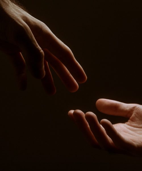 A photograph depicting two hands reaching towards each other, but not touching, in a darkened backdrop
