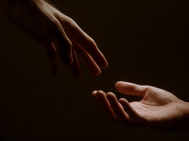 A photograph depicting two hands reaching towards each other, but not touching, in a darkened backdrop