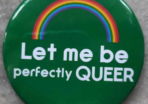 A green pin badge with a rainbow on top and right below it is a text that reads: “Let me be queer” in white font colour.