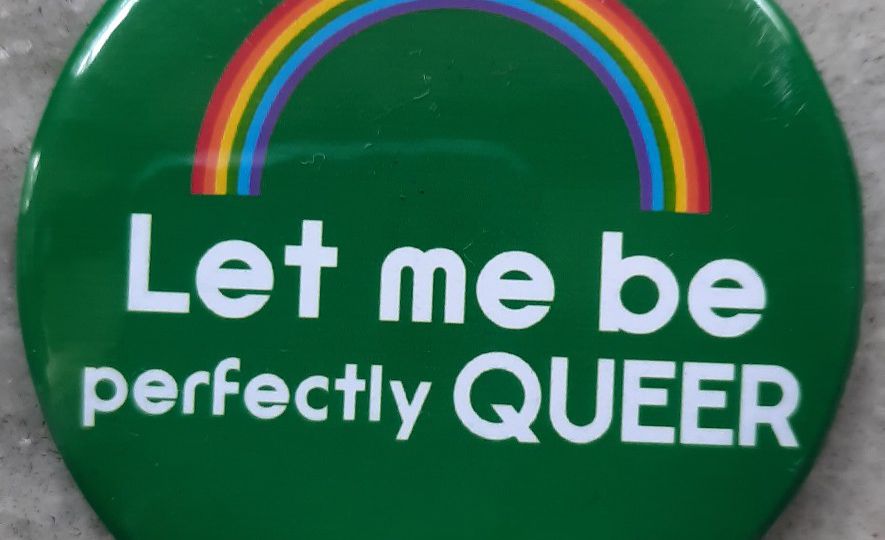 A green pin badge with a rainbow on top and right below it is a text that reads: “Let me be queer” in white font colour.