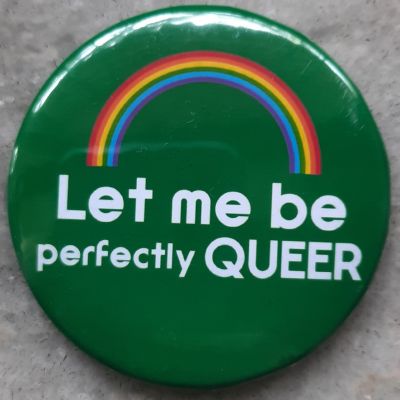 A green pin badge with a rainbow on top and right below it is a text that reads: “Let me be queer” in white font colour.