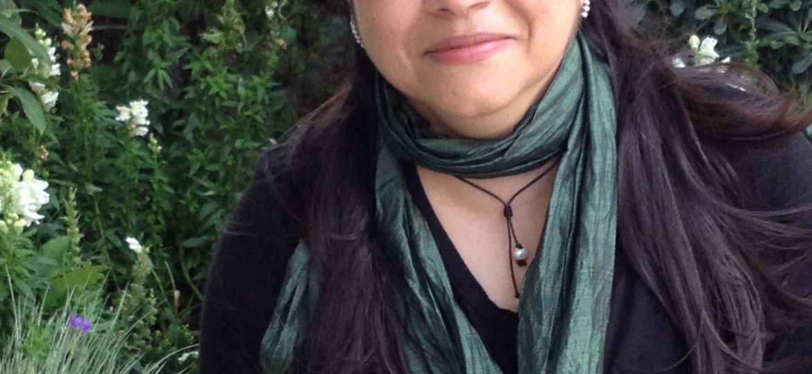 Photo of Sameera Khan. She has long black hair and is standing in a garden. She is wearing a black kurta, a deep emerald dupatta and a black necklace.