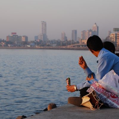 A photo of heterosexual couple sitting cross-legged on a rampart beside the sea. The man is wearing a blue shirt and there are shopping bags beside him. Beside him, only the mid-length hair of the woman can be seen. In the horizon is the city skyline of Mumbai with skyscrapers and other buildings at golden hour.
