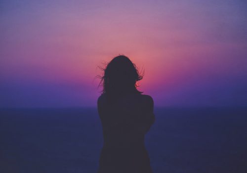 A photograph of the silhouette of a woman with long hair with the horizon of an ocean in the background and a sky at sunset with purple, pink and yellow gradient