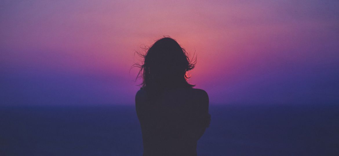 A photograph of the silhouette of a woman with long hair with the horizon of an ocean in the background and a sky at sunset with purple, pink and yellow gradient