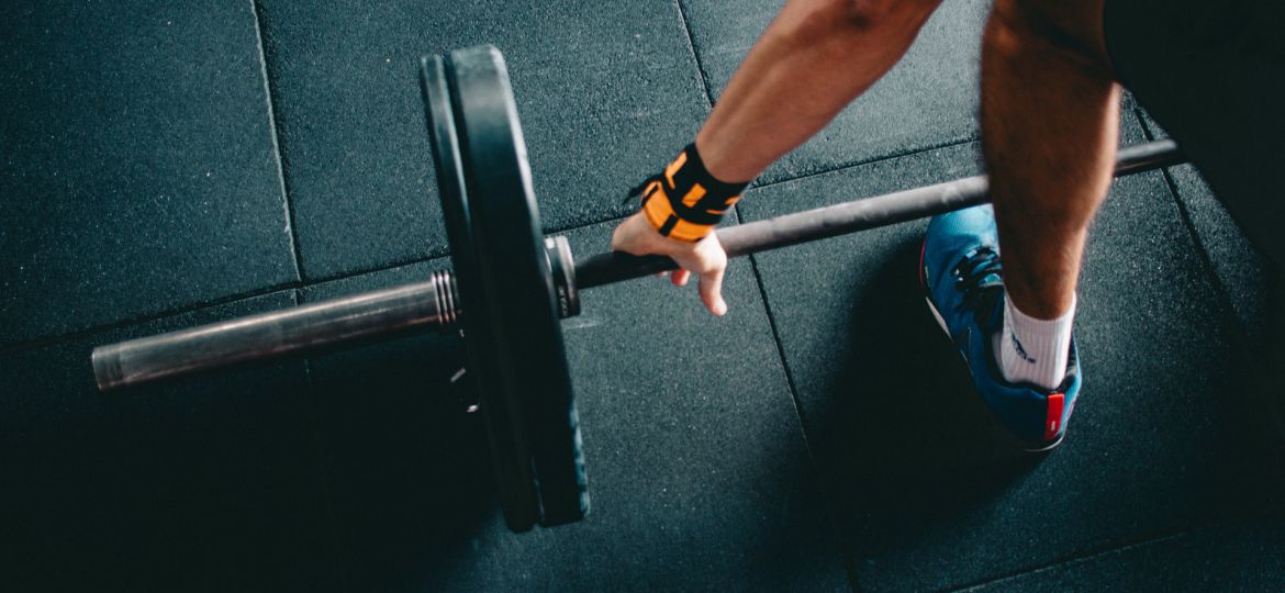 A photograph of a person in a gym holding a barbell, his arm and leg visible in the frame.