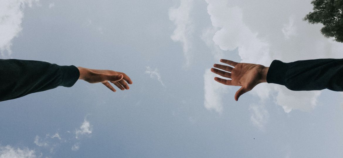 खुले आसमान के तले दो लोग एक दूसरे की तरफ़ अपना हाथ बढ़ाते हुए. Two hands reaching out to each other against the background of a blue sky.