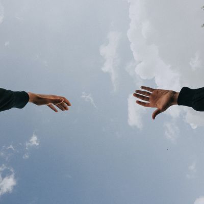खुले आसमान के तले दो लोग एक दूसरे की तरफ़ अपना हाथ बढ़ाते हुए. Two hands reaching out to each other against the background of a blue sky.