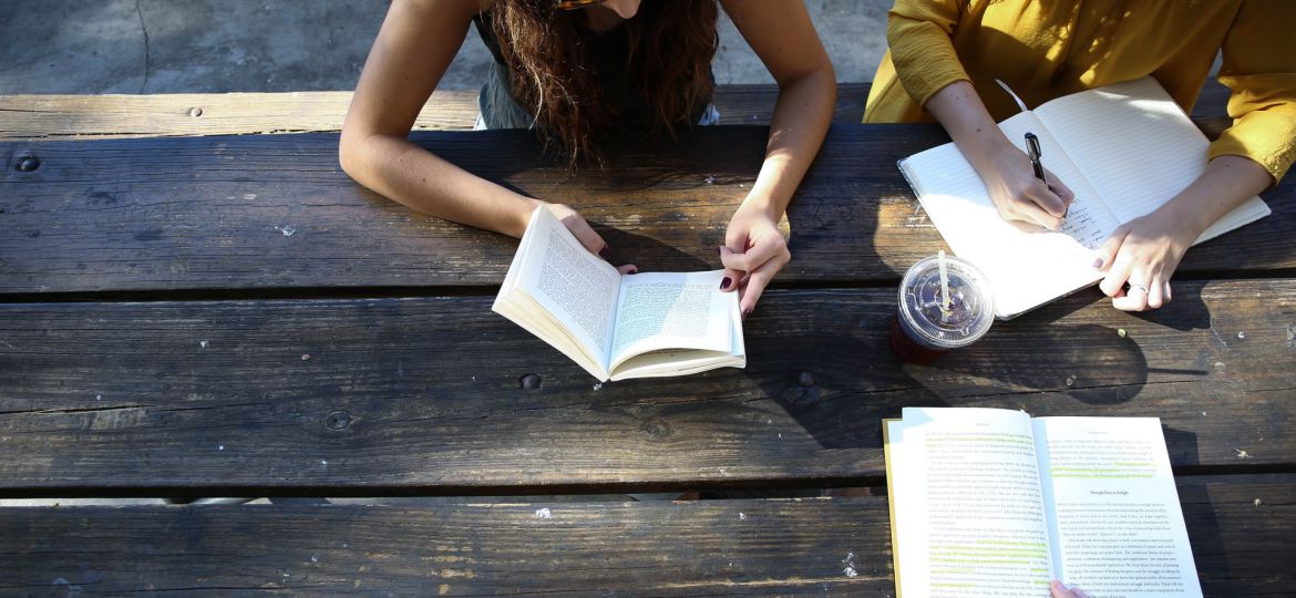 Three people are seated at a wooden table - two are reading and one is writing.