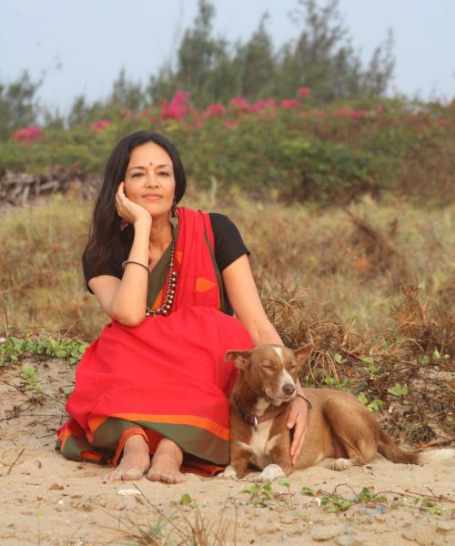 Photograph of a woman with long hair in red sari, sitting with a brown dog against a background of bushes with vines and pink azaleas. The woman is resting her chin on one hand and petting the dog with the other.